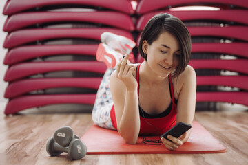 Young woman resting after workout. Healthy young female taking break after training in gym with phone in hands.