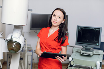 Close up portrait of young caucasian woman doctor in red uniform, holding digital tablet pc, posing near modern ultrasonic lithotripter machine in urology medical center