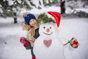 Happy child girl plaing with a snowman on a snowy winter walk