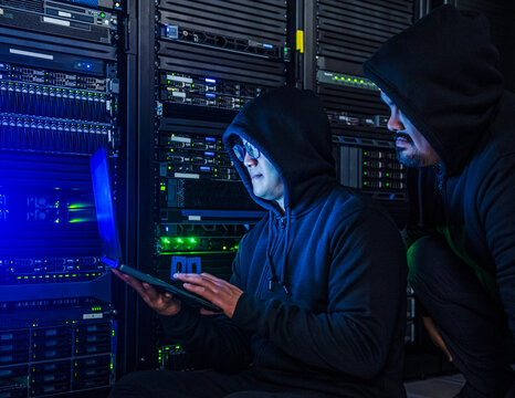 Two men in hooded sweatshirts look at laptop next to server racks