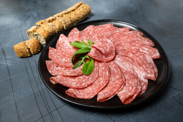 Traditional Italian salami on a black plate served with arugula and multi-grain baguette