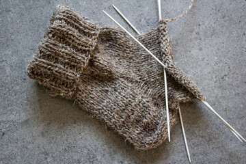 Knitted warm socks made of organic wool. close up photo of brown sock, knitting needles and ball of natural yarn. Grey background with copy space. 