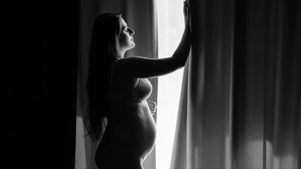 Black and white portrait of beautiful pregnant woman in lingerie looking out of the window at morning