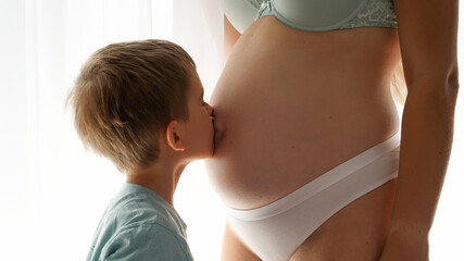 Closeup of little boy kissing his pregnatn mom in big belly standing at window in bedroom. Concept of family happiness and baby anticipation