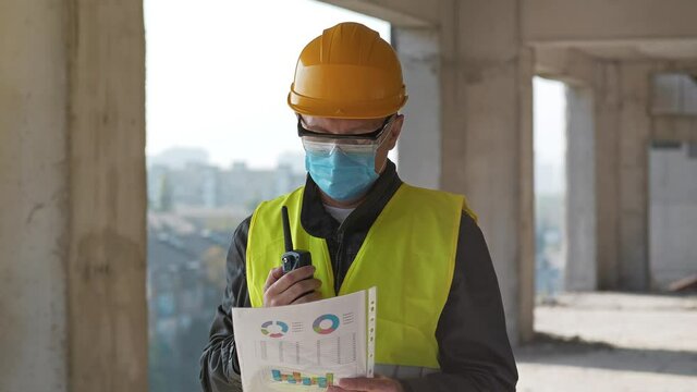 Workman in medical protective mask, goggles and hard hat stands at construction site, holds in hand work papers and speaks via portable radio transmitter
