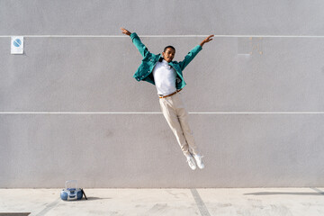 Young man with radio on footpath jumping against gray wall