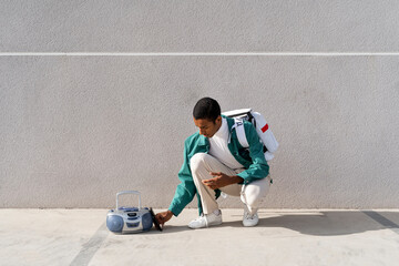 Young man with bag crouching by radio against gray wall