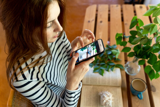 Young woman photographing coffee cup and book on table through mobile phone while sitting at home