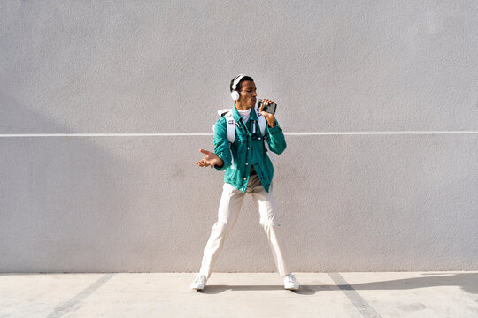 Young man with headphones and backpack singing while standing against gray wall