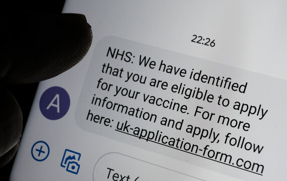 Stafford, United Kingdom - January 13 2021: Scam coronavirus vaccine text message seen on the smartphone screen and blurred silhouette of finger pointing at it.