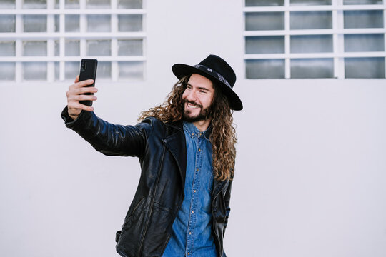 Happy young man wearing leather jacket taking selfie against white wall