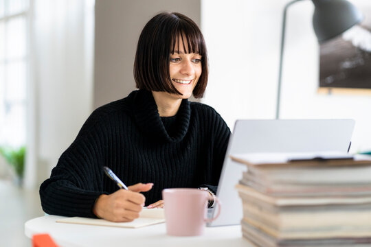 Smiling Young Female Student With Laptop Writing While Studying In Study Room