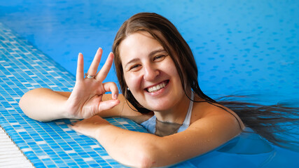 Portrait of happy smiling brunette woman relaxing in swimming pool and showing OK sign with fingers.