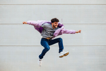 Carefree young man with arms outstretched jumping against gray wall