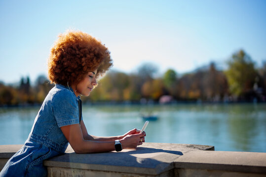 Mid adult woman with frizzy hair using mobile phone at Estanque Grande Del Retiro pond on sunny day, Spain