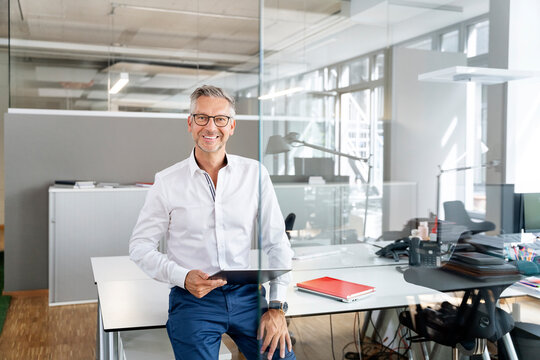 Happy businessman with digital tablet leaning on desk in office