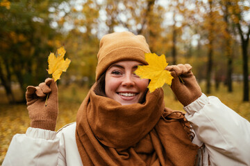 Happy young woman covering eye with autumn leaf in public park