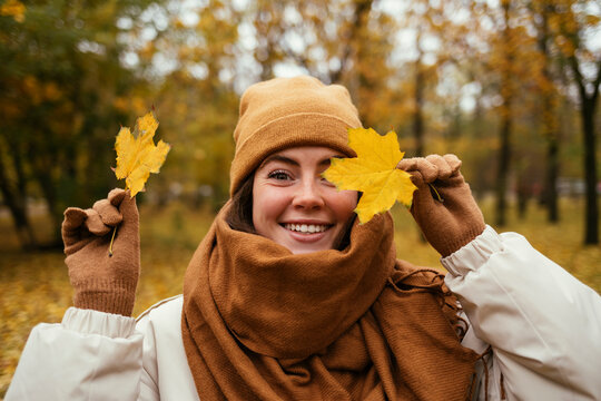 Happy young woman covering eye with autumn leaf in public park