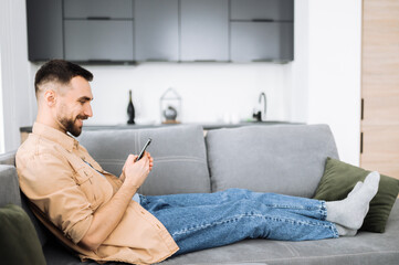 Happy caucasian man sits at the couch, using smartphone. Handsome guy got a dream job or a good news or message. Attractive stylish guy chatting online with friends or family