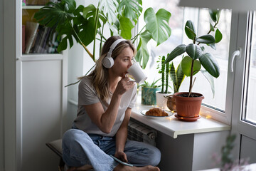 Woman listening to music wear wireless headphones, sitting and looking out the window, drinking tea...