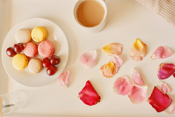 Breakfast in bed with macaroons. Cup of fragrant coffee, saucer with dessert and cherries on table. Rose petals on white tray. Surprise for Valentine's day. Top view.