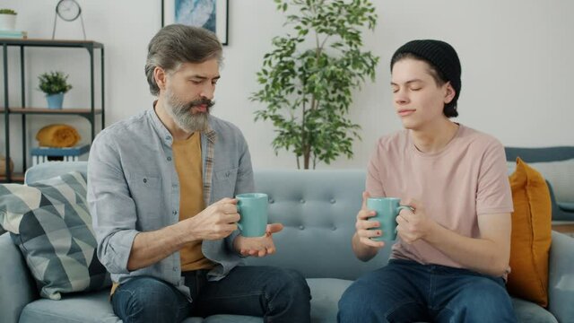 Caring kid is bringing drink for dad enjoying tea and talking sitting on couch together in apartment. Father and son relations and leisure time concept.