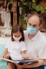 Stock Photo - Little granddaughter in a face mask with grandfather are self-educating on self-isolation.  Family support during quarantine isolation due to outbreaks of coronavirus. Elderly at risk