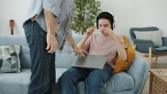 Kid is enjoying music and using laptop when angry dad is coming turning off device arguing with teenager. Modern technology and adolescence concept.