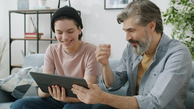 Slow motion of teenage boy and father using tablet chatting then doing high-five hand gesture and laughing enjoying family activity and device