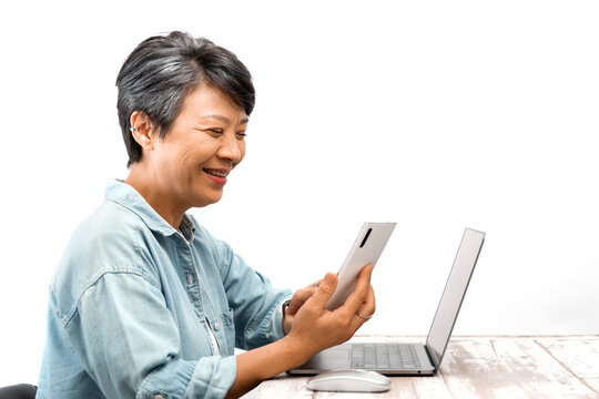 Happily active Senior businesswoman working on a laptop while looking at a cellphone in office. Asian mature female working on a laptop computer and sitting at her desk. Isolated white background.