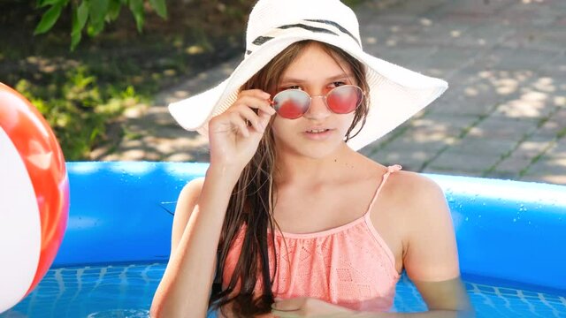 Beautiful smiling teenage girl in summer hat and stylish sunglasses relaxing in inflatable swimming pool at house backyard. Concept of happy summer holiday and vacation