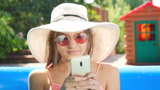 Portrait of smiling young girl in hat and sunglasses relaxing in swimming pool and using smartphone. Concept of happy summer holidays and vacation