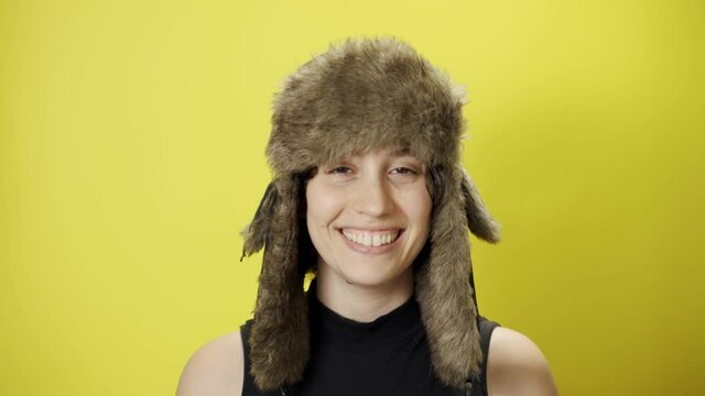 Happy young woman with fur trapper winter hat looks and smiles at camera. Cold weather conditions or fashion shoot. Beautiful and attractive female with fun and joyful look on yellow background