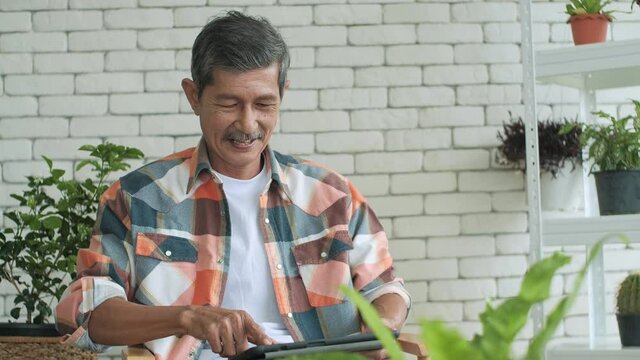 Elderly Asian man relaxing holding digital tablet reading e book in garden at home, retirement man concept. senior adult man enjoying using computer pad apps browsing internet shopping.