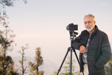 A SENIOR ADULT MAN STANDING WITH CAMERA ON A HILLTOP	