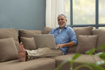 A HAPPY OLD MAN SITTING COMFORTABLY ON SOFA WITH LAPTOP 