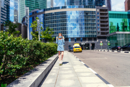 slender girl teenager in denim clothes and a medical mask rides a skateboard around the city on a sunny summer day