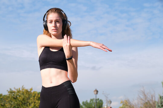 Redhead caucasian woman with headphones stretching her arm. Fitness and sport outdoor.
