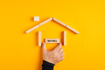 Fototapeta na wymiar Male hand placing a buy or sell sign in a house made of wooden blocks on yellow background.