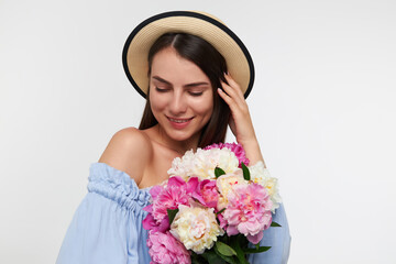 Young lady, pretty woman with long brunette hair. Wearing a hat and blue dress. Holding bouquet of flowers and touching her hair. Watching down, isolated over white background