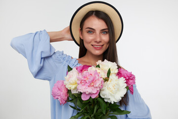 Teenage girl, happy looking woman with brunette long hair. Wearing a hat and blue dress. Holding bouquet of flowers and touching her head. Watching at the camera isolated over white background
