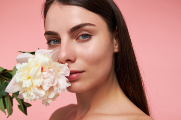 Nice looking woman, beautiful girl with long brunette hair and healthy skin, holding a flower next to her face. Watching at the camera, closeup, isolated over pastel pink background