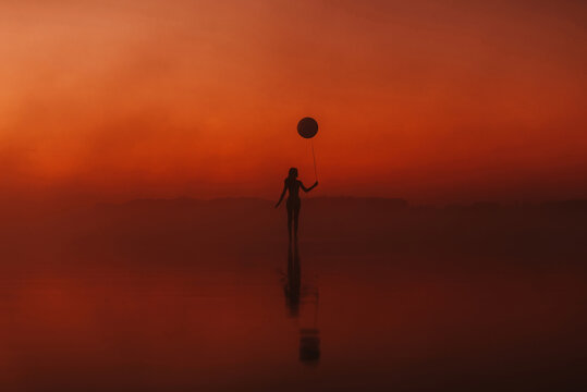 surreal silhouette of a girl with a balloon in her hand on the water at sunrise in the fog in summer
