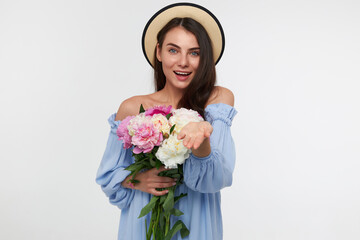 Teenage girl, happy looking woman with brunette long hair. Wearing a hat and blue dress. Holding a bouquet of flowers and showing open palm. Watching at the camera isolated over white background