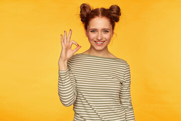 Teenage girl, happy, successful looking red hair woman with two buns. Wearing striped sweater and showing okay sign, smile. Watching at the camera isolated over yellow background