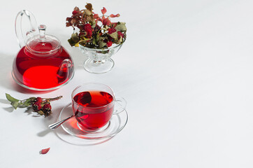 Autumn teatime with red herbal tea in transparent tea pot, cup with hawthorn berries, rose hip, dry leaves in sunlight on white wood table, copy space.