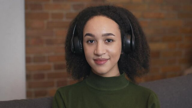 Close up portrait of young african american woman wearing wireless headset, enjoying audiobook, smiling to camera
