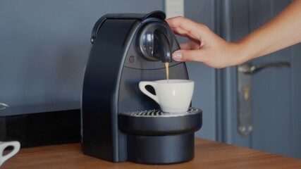 A caucasian woman's hand is pushing a button on the capsule coffee machine to extract espresso in a white ceramic cup. Grey wall in the background. High-resolution jpg photo - 405418563