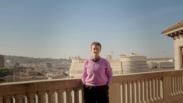A caucasian young lady with a short haircut, glasses in a purple sweatshirt is standing on the balcony with lowered coronavirus black face mask. Barcelona view. High-quality cowboy shot jpg portrait