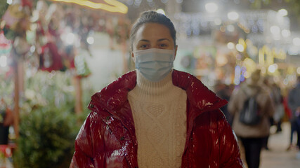 Brunette young woman in a red shiny puffer coat standing in a covid face mask in the middle of a Christmas market. Blurred winter festive lights on the background. Medium shot front portrait.